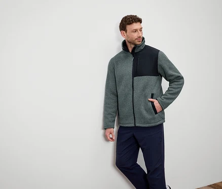 Ein Mann lehnt an einer Wand und trägt eine graue Fleecejacke mit schwarzen Schultern und dunkelblaue Hosen.