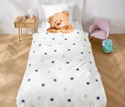 Im Zimmer steht ein Bett mit weißer Bettwäsche mit blauen Punkten und einem Teddybär. Über dem Bett hängt eine Girlande aus lila Dreiecken.