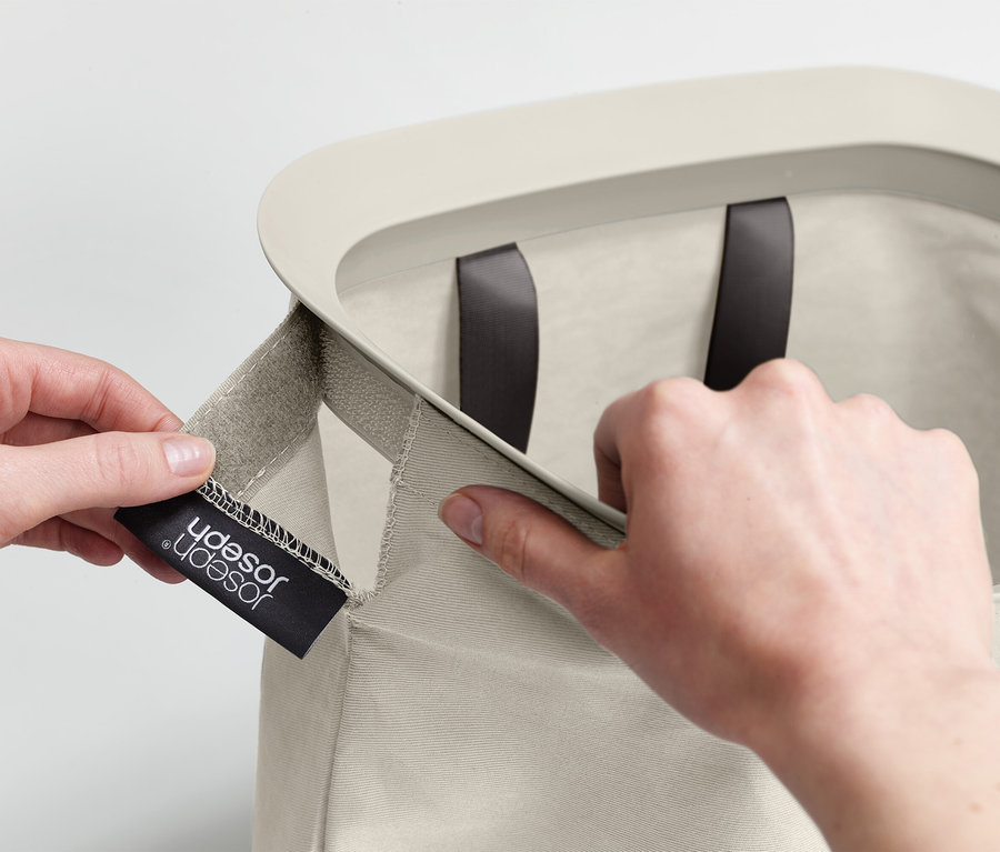 Hände halten einen beigen Stoffkorb mit einem schwarzen Joseph Joseph-Etikett.