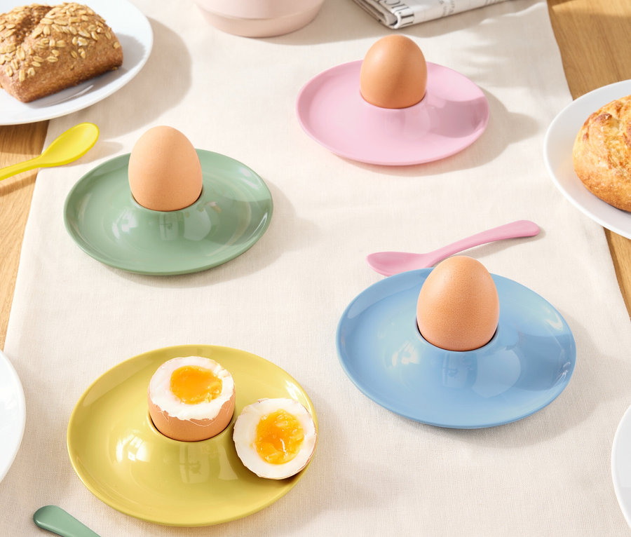 Verschiedenfarbige Eierbecher mit Eiern und Löffeln auf einer Tischdecke.
