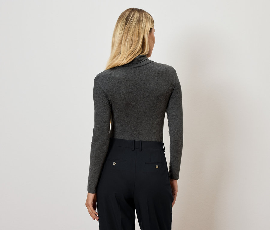 Rückansicht einer Frau mit blonden Haaren in einem grauen Rollkragenpullover und einer schwarzen Hose.