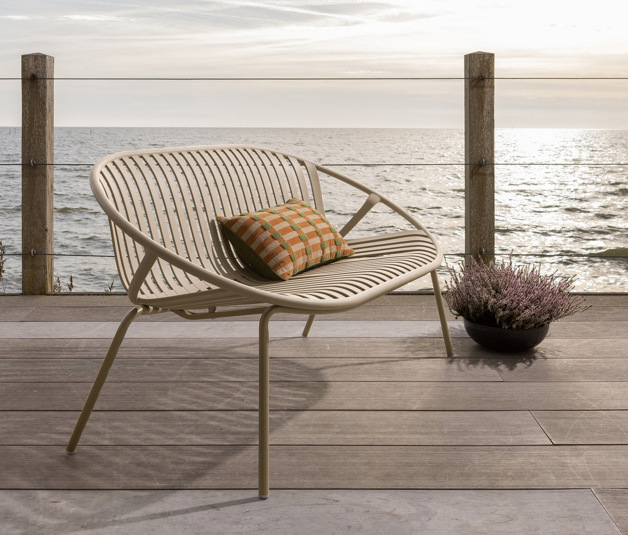Ein beiger Stuhl mit einem karierten Kissen steht auf einer Holzterrasse mit Blick auf das Meer.