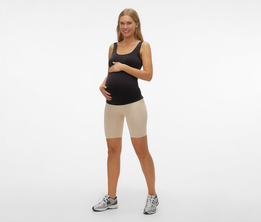 Schwangere Frau mit schwarzem Tanktop und MAMALICIOUS Skinny Shorts »MLTIA JEANNE« in Schwarz posiert vor einem weißen Hintergrund.