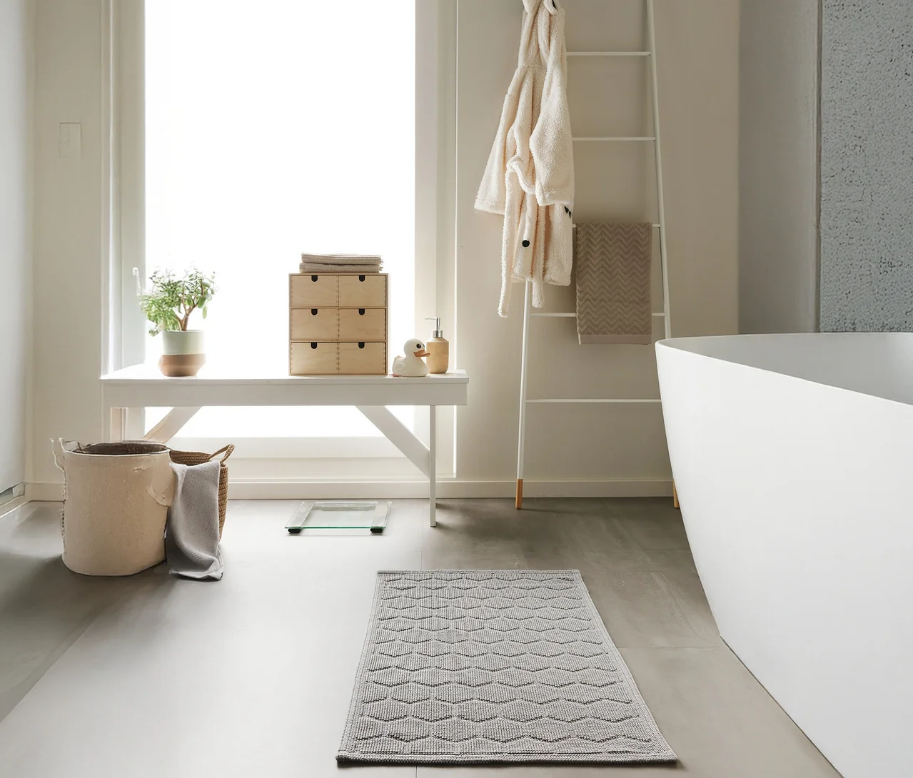 Ein Badezimmer mit einer weißen Bank, einer Leiter und einer Badewanne. Auf dem Boden liegt eine hellgraue Badematte »Jojo« von benuta mit Rautenmuster (50 x 80 cm).
