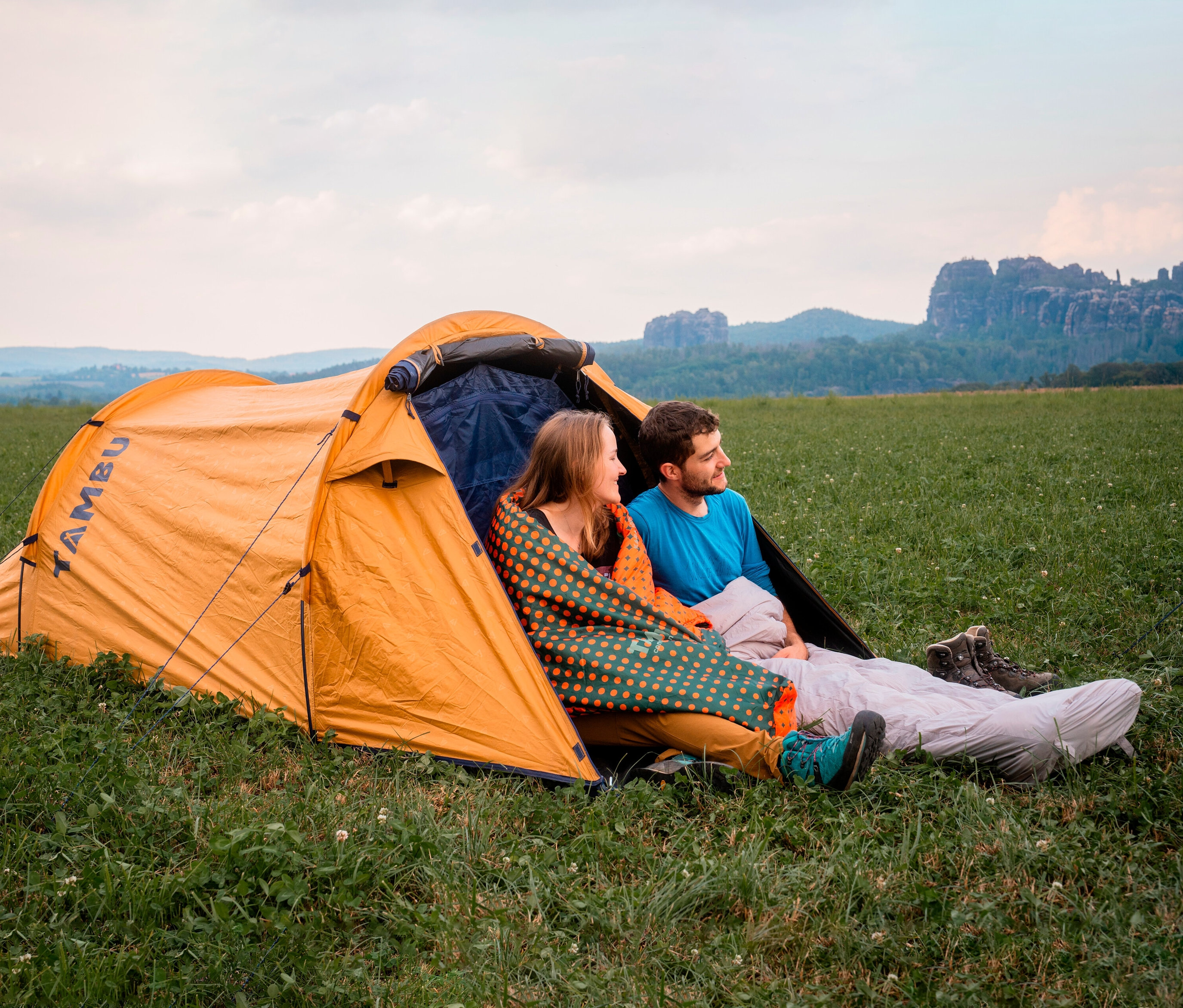 Ein Paar sitzt im geöffneten, orangefarbenen Zelt, die Frau ist in einen »KARA 1.000« Mumienschlafsack von Tambu gehüllt.