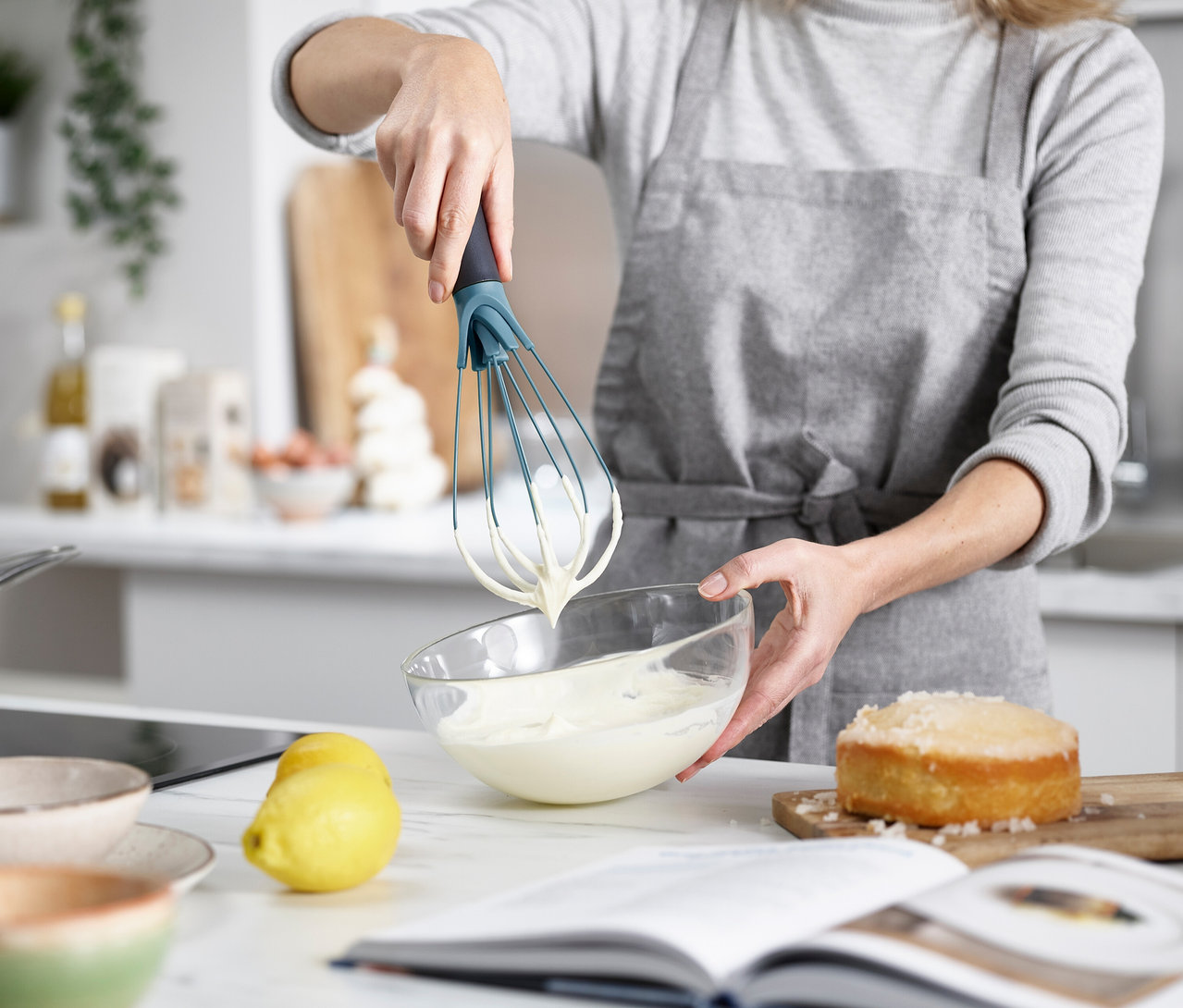 Eine Frau verrührt mit einem Schneebesen Teig in einer Glasschüssel, während ein Kuchen und eine Zitrone auf der Küchentheke stehen.