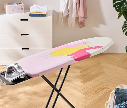 Ein Bügeleisen steht auf einem Bügelbrett mit einem rosa Bezug mit gelben und pinken Lamas. Im Hintergrund befinden sich eine Kommode, Wäsche und eine Zimmerpflanze.