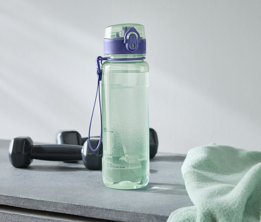 Eine grüne Wasserflasche mit lila Deckel steht neben zwei schwarzen Hanteln und einem hellgrünen Handtuch auf einem grauen Untergrund.