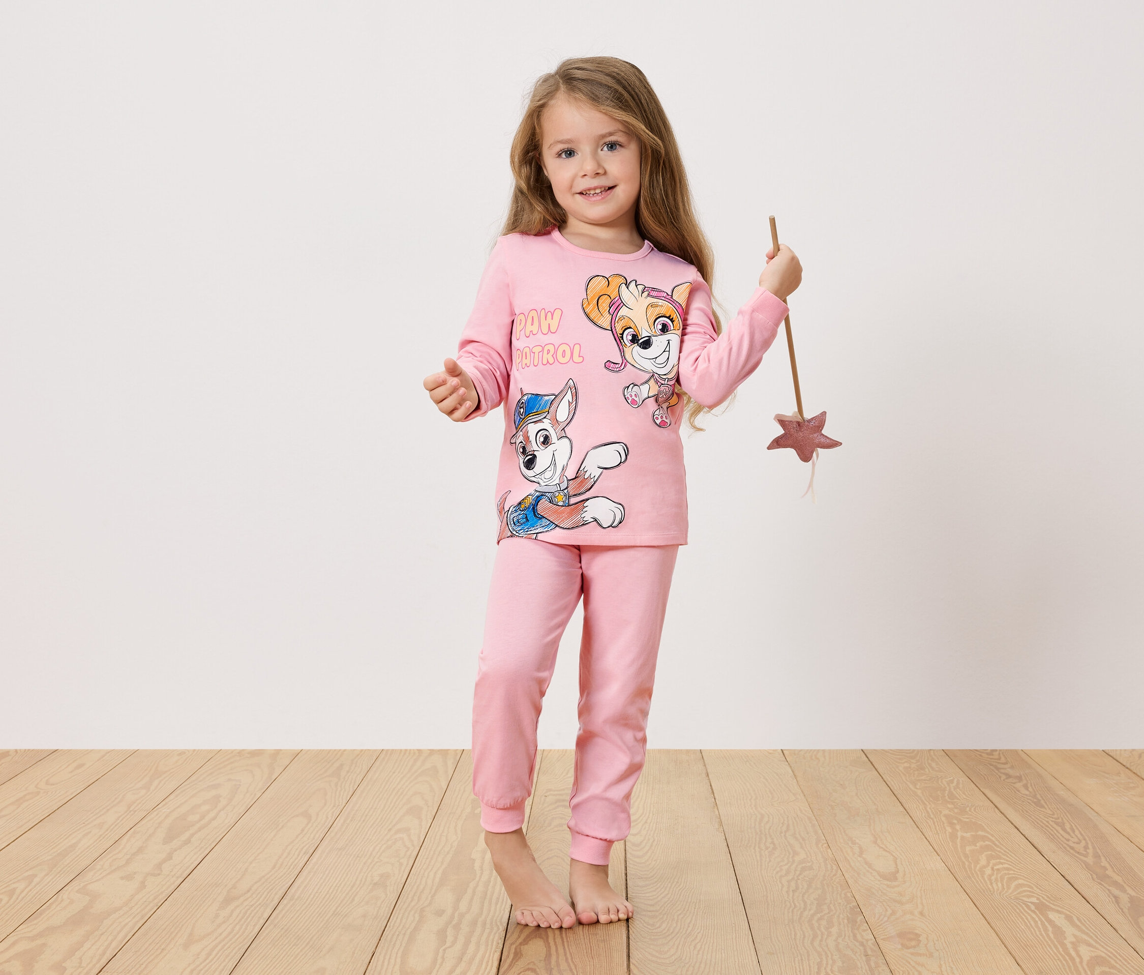 Mädchen mit langem blondem Haar trägt einen rosa Paw Patrol Pyjama und hält einen Zauberstab in der Hand.