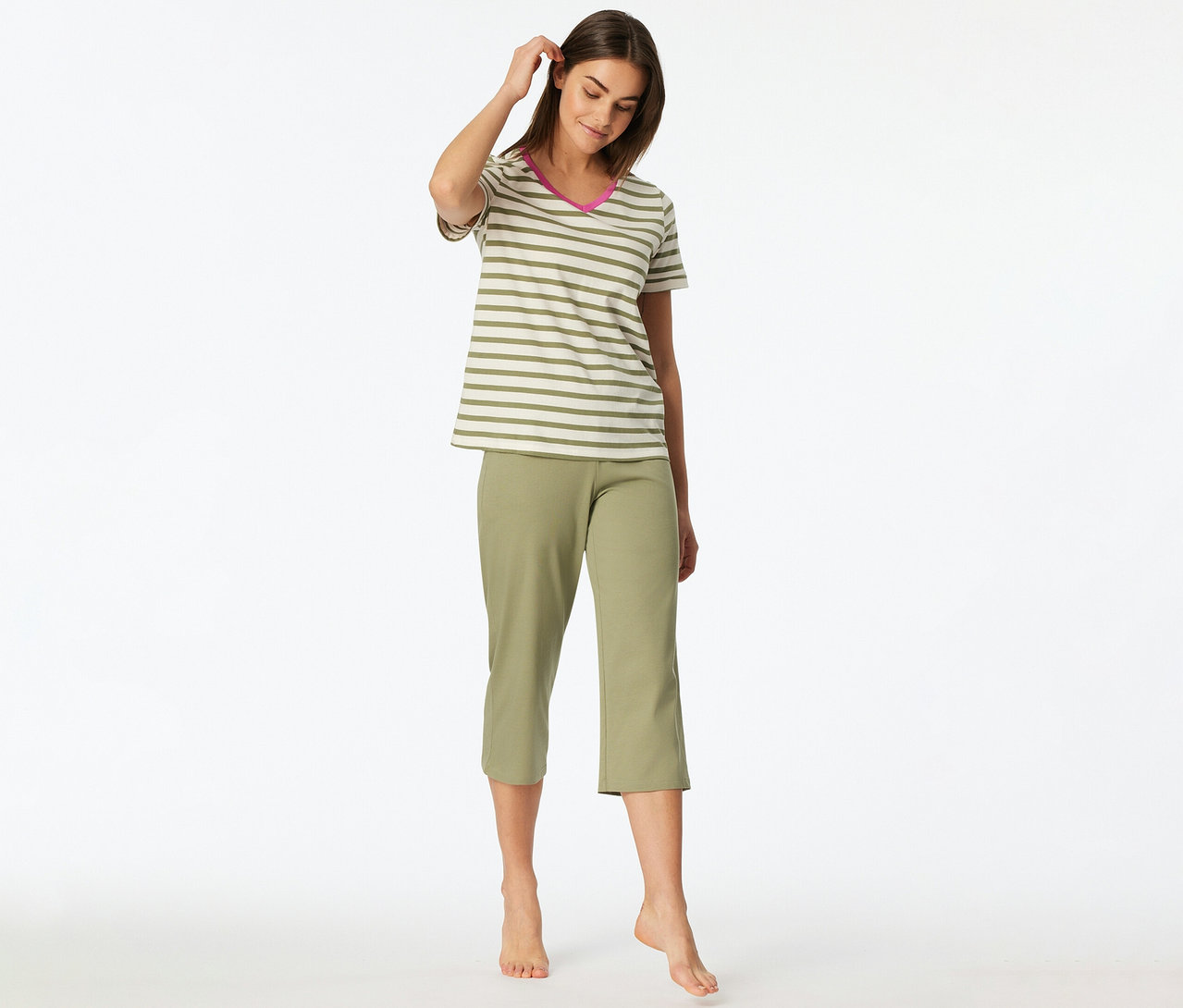 Eine Frau steht barfuß, blickt nach unten und berührt ihr Haar, bekleidet mit einem SCHIESSER 3/4-Pyjama-Set, bestehend aus einem gestreiften Kurzarm-Oberteil mit pinkem V-Ausschnitt und einer olivgrünen 3/4-Hose.