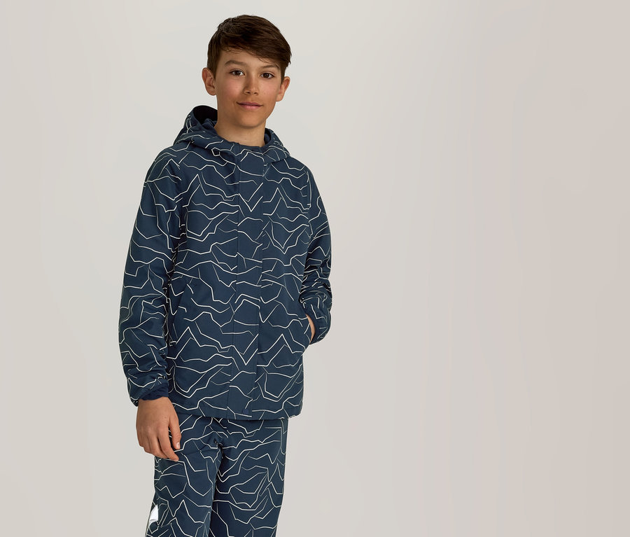 Ein Junge steht mit einer Hand in der Tasche in einem marineblauen Hoodie und einer Hose mit weißem Zickzackmuster.