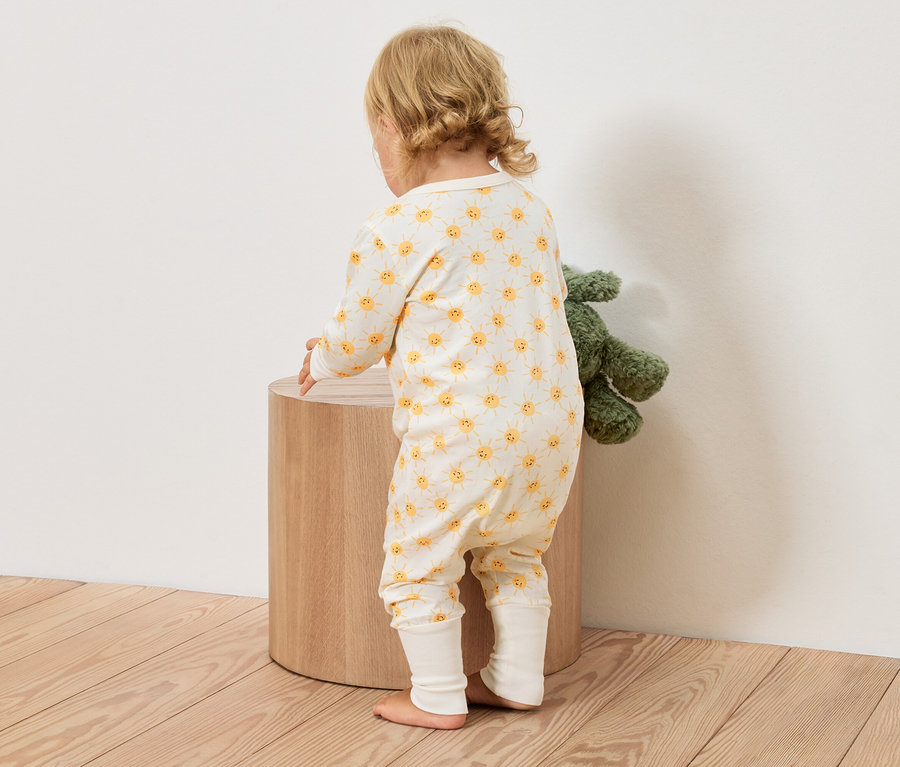 Kleinkind in einem gelb-weissen Overall mit Sonnenmuster steht neben einem runden Holzhocker, ein grünes Stofftier daneben.