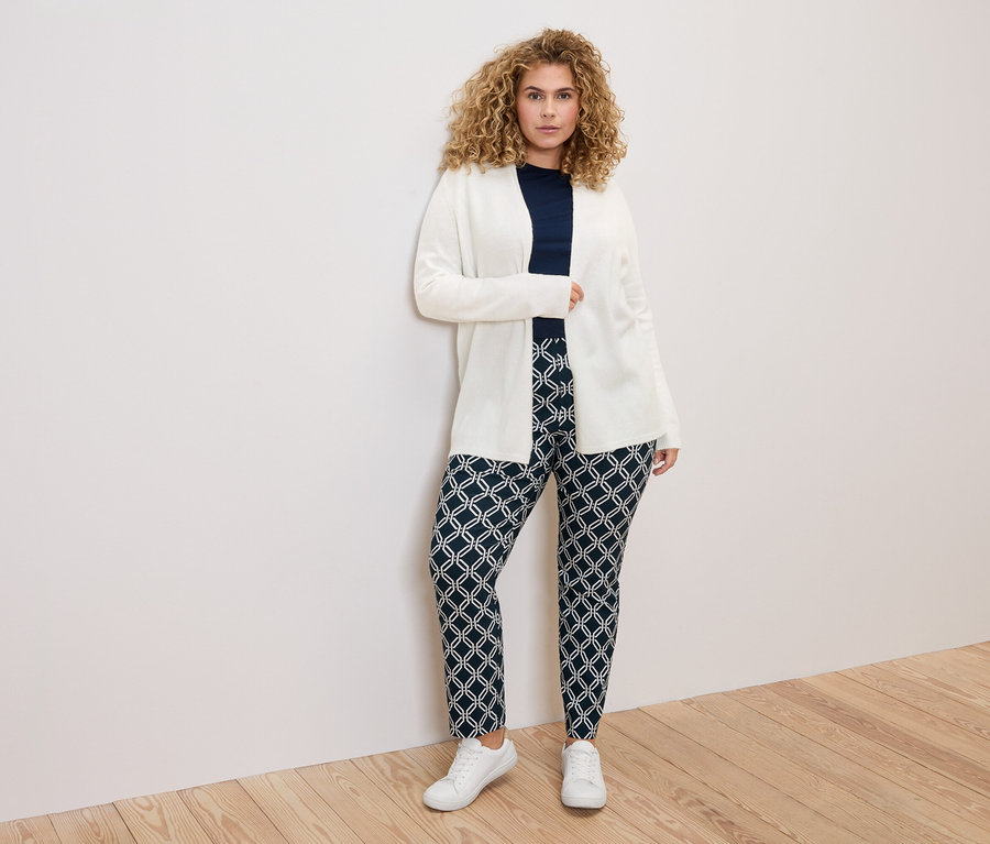 Frau mit lockigem Haar, die eine weiße Strickjacke, ein marineblaues Oberteil und gemusterte Hosen vor einer weißen Wand trägt.