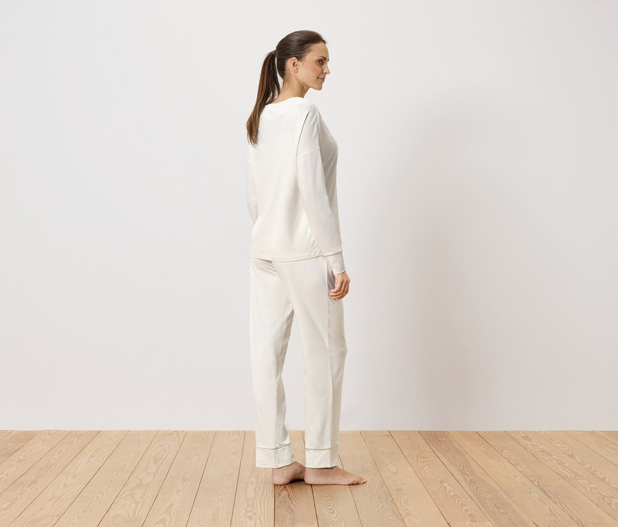 Frau steht seitlich in cremefarbenem, langärmligem Pyjama vor einer weißen Wand.