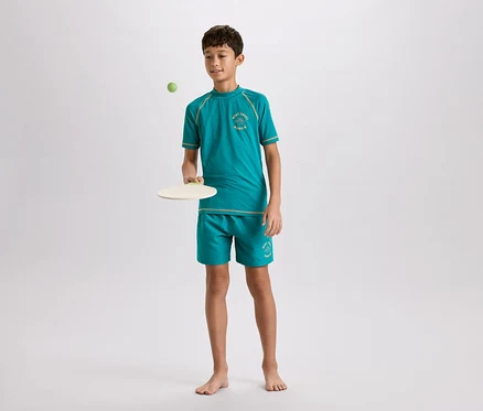Ein junger, barfüßiger Junge in einem türkisfarbenen T-Shirt und Shorts mit hellbraunen Akzenten spielt mit einem Paddelball vor einem hellgrauen Hintergrund.