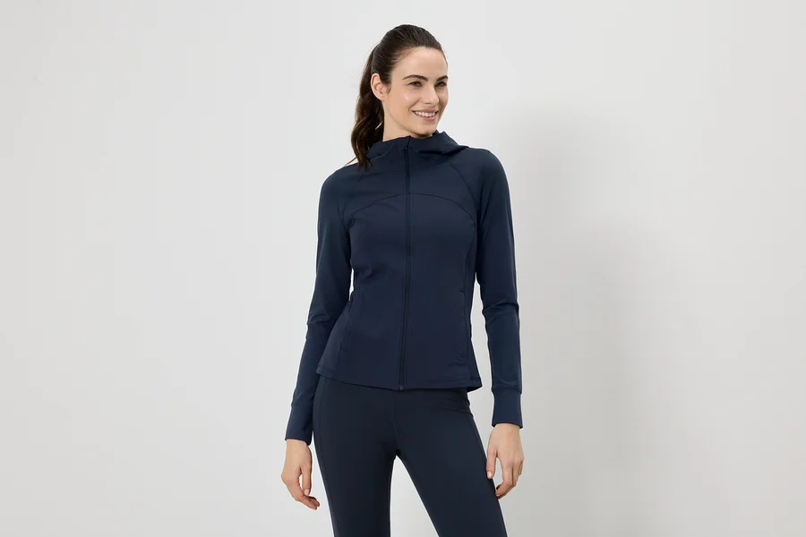 Frau mit Pferdeschwanz trägt marineblaue Jacke mit Kapuze und Leggings vor einem weißen Hintergrund.