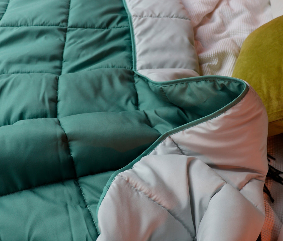 Nahaufnahme einer aufgeschlagenen, gesteppten, mintgrünen Decke mit hellgrauer Innenseite auf einem Bett mit Kissen und einer weißen Decke.