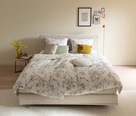 Ein gemachtes Bett mit der SCHÖNER WOHNEN-Kollektion Bettwäsche »LOVELY« mit floralem Muster in Gelb, Orange und Grau. Links steht ein Nachttisch mit gelben Blumen in einer Vase, rechts eine goldene Stehlampe.