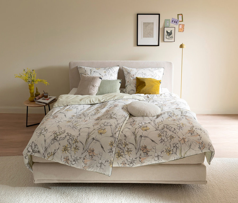 Ein gemachtes Bett mit der SCHÖNER WOHNEN-Kollektion Bettwäsche »LOVELY« mit floralem Muster in Gelb, Orange und Grau. Links steht ein Nachttisch mit gelben Blumen in einer Vase, rechts eine goldene Stehlampe.