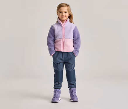 Mädchen mit lila-rosa Fleecejacke, blauen Hosen und lila Stiefeln steht vor hellem Hintergrund.