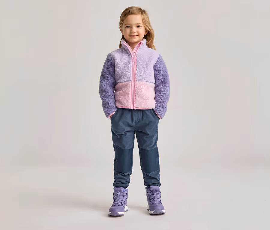 Mädchen mit lila-rosa Fleecejacke, blauen Hosen und lila Stiefeln steht vor hellem Hintergrund.