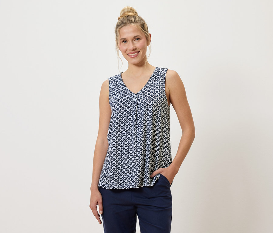 Frau mit Dutt trägt ein blau-weiß gemustertes Top und blaue Hose, eine Hand in der Hosentasche.