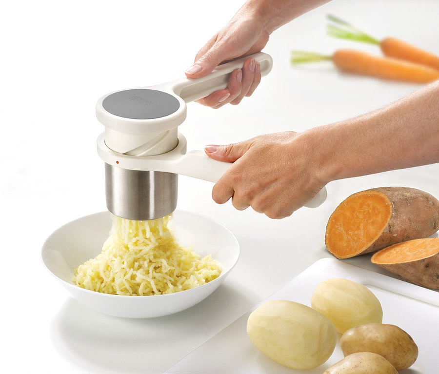 Hände benutzen ein Joseph Joseph Pressen-Set, 3-teilig, um Kartoffeln in eine Schüssel zu pressen. Süßkartoffeln und Karotten liegen daneben.