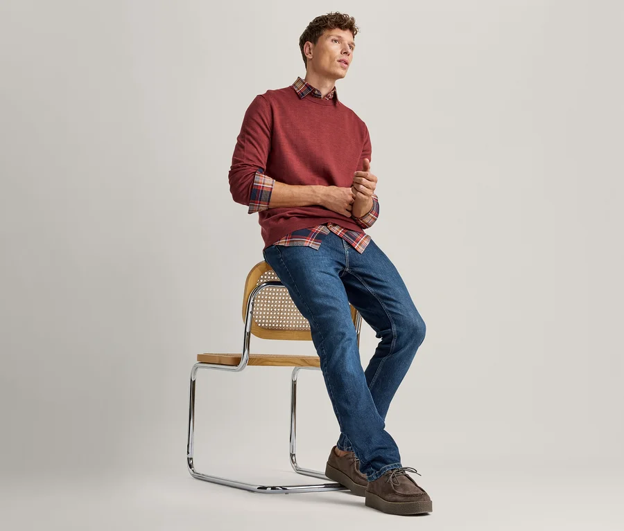 Junger Mann sitzt auf einem Stuhl mit Holzgeflecht und Chromgestell, trägt einen roten Pullover über einem karierten Hemd, blaue Jeans und braune Schuhe. Er blickt nach rechts.