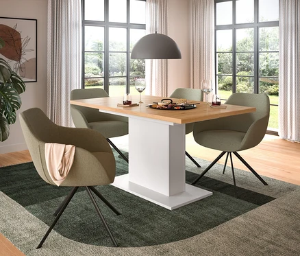 Ein modernes Esszimmer mit hellem Holztisch, weißem Säulenfuß und vier olivgrünen Stühlen. Auf dem Tisch stehen eine Servierplatte und Weingläser. Eine Monstera-Pflanze, ein geometrischer Teppich und eine graue Pendelleuchte schmücken den Raum.