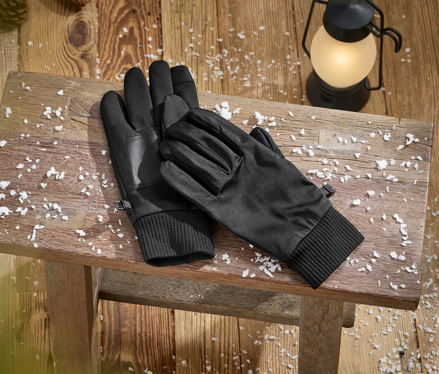 Ein Paar schwarze Handschuhe mit gerippten Bündchen und strukturierten Handflächen liegt auf einer rustikalen Holzbank, bedeckt mit Kunstschnee. Eine leuchtende Laterne und Tannenzapfen sind im Hintergrund auf dem Holzboden zu sehen.