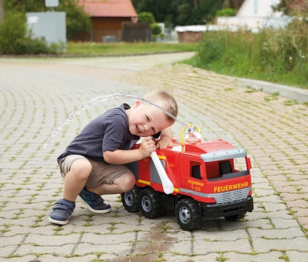 Ein lächelnder Junge hockt neben einem roten GIGA TRUCKS Feuerwehr »Actros« Spielzeug-Feuerwehrauto, das Wasser über seinen Kopf spritzt.