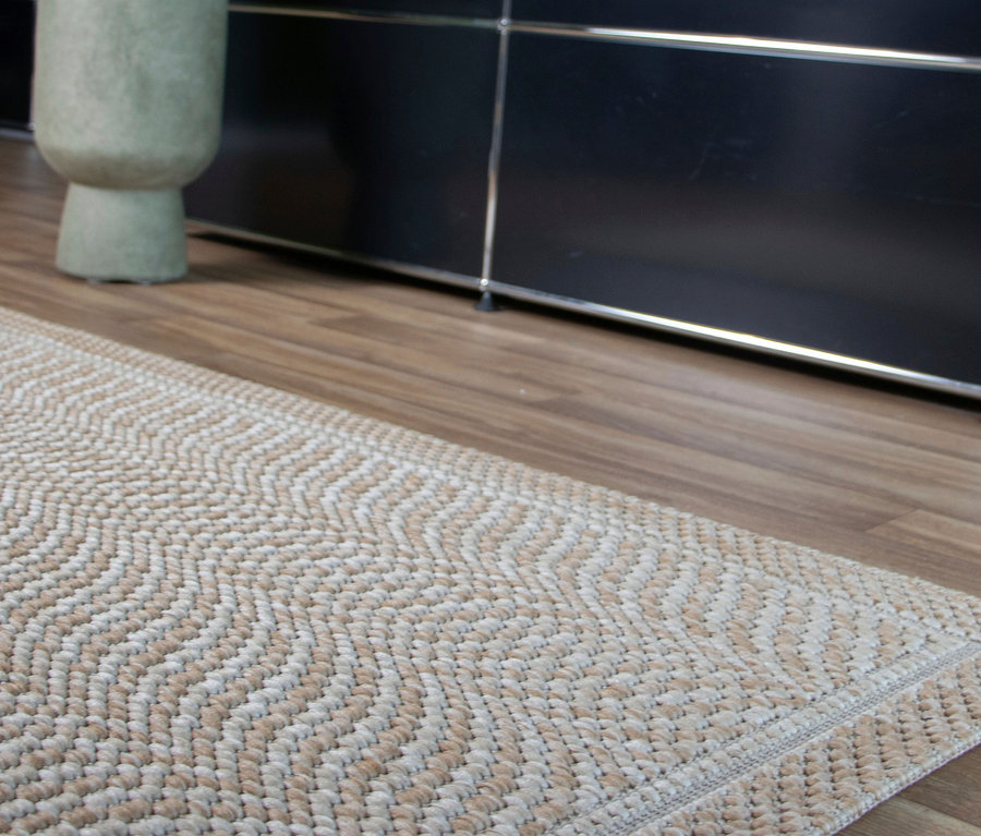 Ein Teppich »Twine« mit beigem und weißem Fischgrätmuster liegt auf einem Holzboden. Im Hintergrund ist ein dunkler Schrank und links ein heller Krug zu sehen.