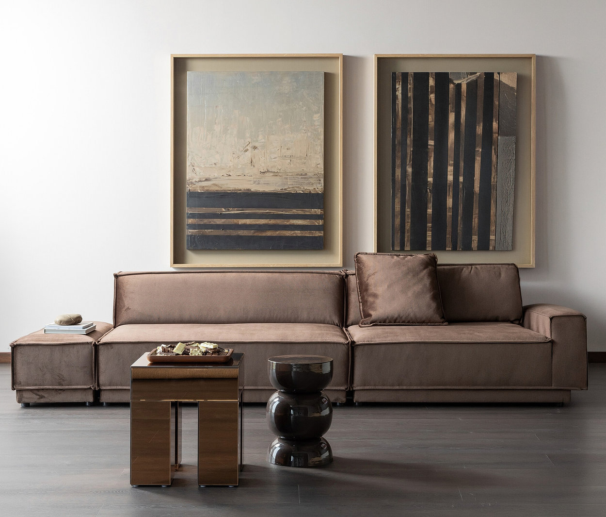 Ein modernes Wohnzimmer mit einem modularen braunen Sofa und Ottoman vor einer weißen Wand. Zwei gerahmte abstrakte Kunstwerke hängen über dem Sofa. Vor dem Sofa stehen ein reflektierender rechteckiger Beistelltisch mit einem Dekotablett und ein dunkler, sanduhrförmiger Hocker auf einem dunklen Holzboden.