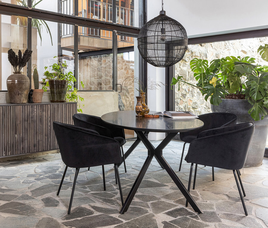 Ein Essbereich mit einem runden schwarzen Tisch und vier schwarzen Samtstühlen auf einem Steinboden. Über dem Tisch hängt eine große schwarze Hängelampe. Links steht ein dunkles Sideboard mit Pflanzen, rechts eine große Monstera.
