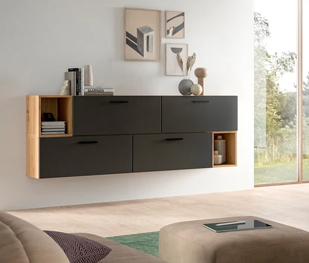 Ein modernes Wohnzimmer mit einem dunkelgrauen, wandmontierten Schrank, der vier geschlossene Fächer und offene Holzregale an den Enden hat. Bücher, Vasen und abstrakte Kunstdrucke schmücken die Wand darüber. Im Vordergrund steht ein hellbrauner Ottoman mit einem Tablet, rechts ist ein großes Fenster mit Blick ins Grüne.