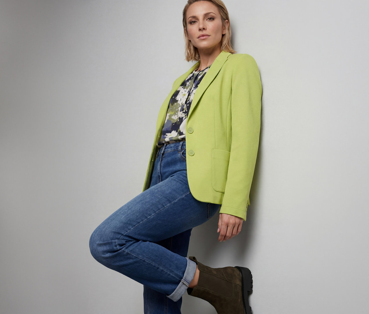 Frau in einem limettengrünen Blazer über einem gemusterten Oberteil, blauen Jeans und dunkelgrünen Stiefeln, die mit einem angewinkelten Bein an einer hellgrauen Wand lehnt.