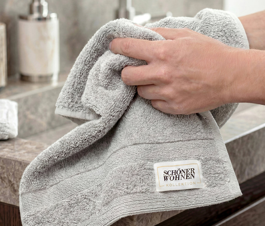 Hände halten ein hellgraues SCHÖNER WOHNEN-Kollektion Gästehandtuch «Cuddly» über einem Waschbecken.