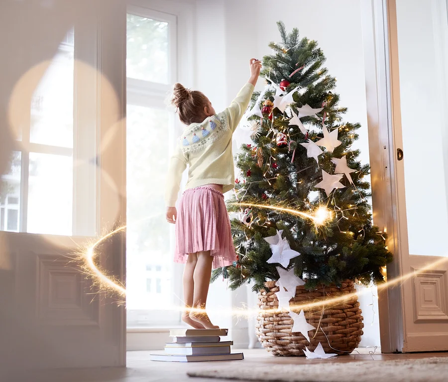 Mädchen steht auf Büchern und schmückt einen Weihnachtsbaum. Sie trägt einen Tüllrock und einen Strickpullover.