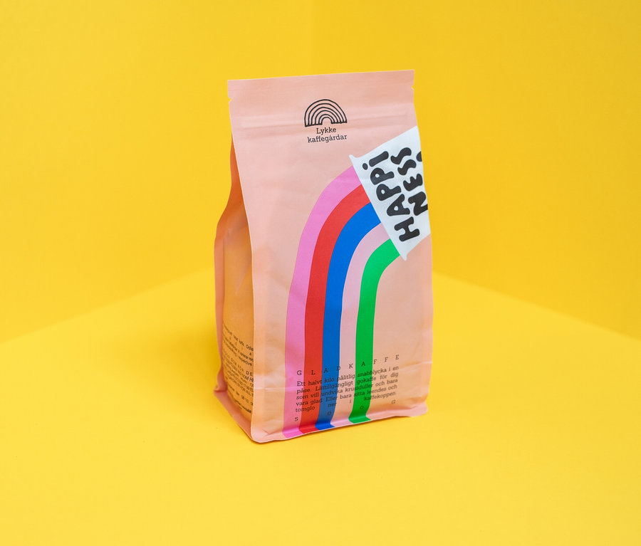 Eine hellrosa Kaffeepackung steht auf einem gelben Untergrund. Auf der Packung sind bunte Streifen und der Schriftzug "Happiness" zu sehen.