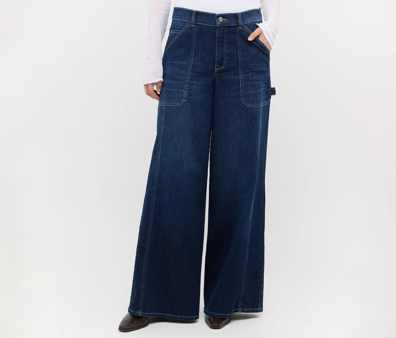 Person in dunkelblauer Wide-Leg-Jeans mit großen Vordertaschen und einer Seitenschlaufe, weißem Langarmshirt und dunklen spitzen Schuhen, eine Hand in der Tasche.