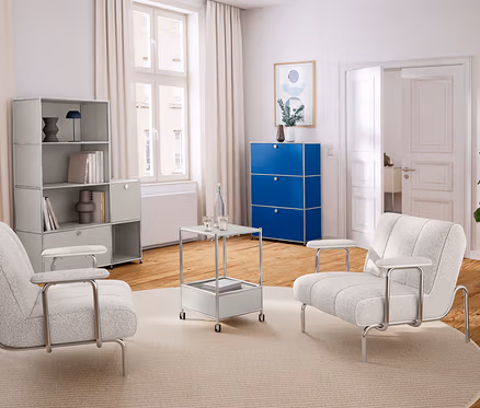 Helles Wohnzimmer mit Holzboden und weißer Einrichtung. Links ein hellgraues Regal, in der Mitte zwei helle Sessel und ein Beistelltisch auf einem runden Teppich. Rechts ein blauer Schrank und eine Pflanze.