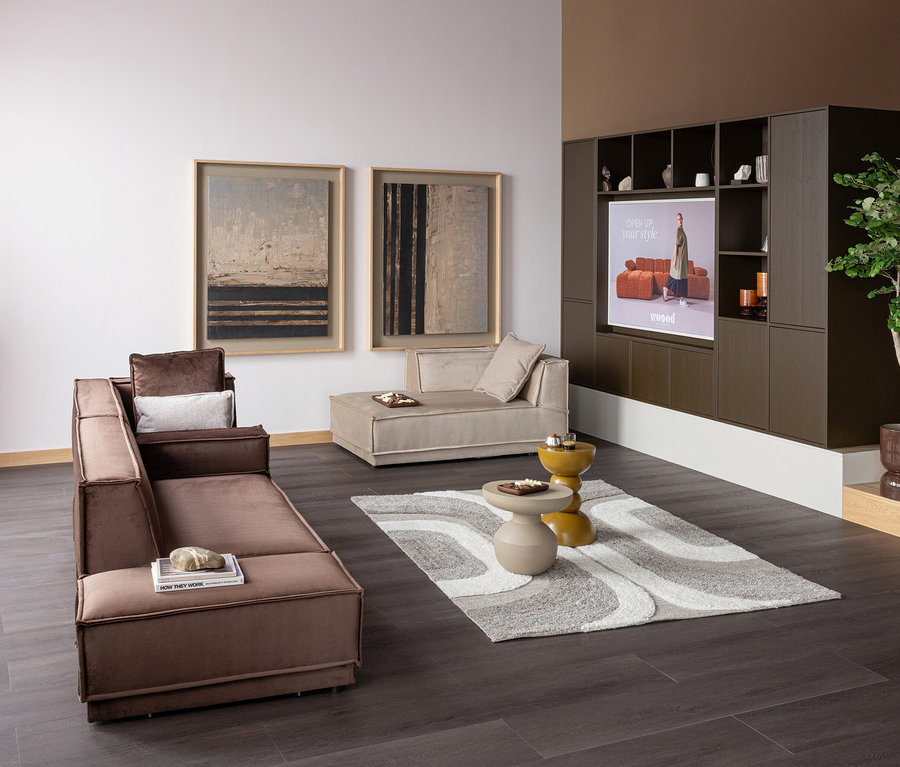 Modernes Wohnzimmer mit braunem L-förmigem Sofa, beiger Chaiselongue und dem grau-weiß gemusterten WOOOD Teppich »Moro«. Auf dem Teppich stehen zwei runde Couchtische, an der rechten Wand eine dunkelbraune Medienwand mit Bildschirm. An der linken Wand hängen zwei abstrakte Bilder.