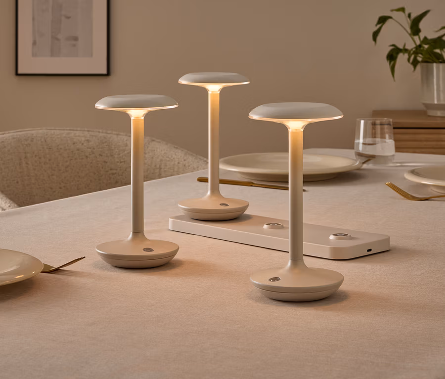 Drei minimalistische, cremefarbene Tischlampen auf einem gedeckten Esstisch, eine davon auf einer Ladestation. Die Lampen leuchten warm.