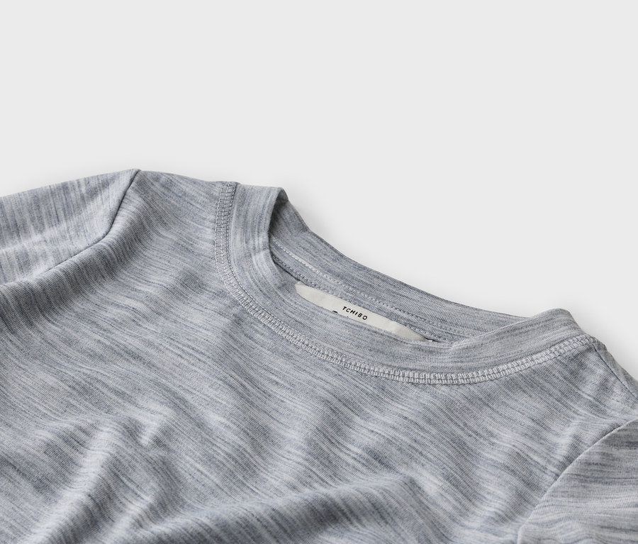 Nahaufnahme eines grauen Melange-T-Shirts mit Rundhalsausschnitt, sichtbaren weißen Nähten am Kragen und einem weißen Etikett mit der Aufschrift „TCHIBO“.