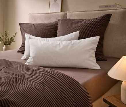 Ein Bett mit beigem Kopfteil, braun gestreifter Bettwäsche und weißen Kissen. Eine Lampe steht auf einem Nachttisch, eine Vase mit Grünpflanzen links.