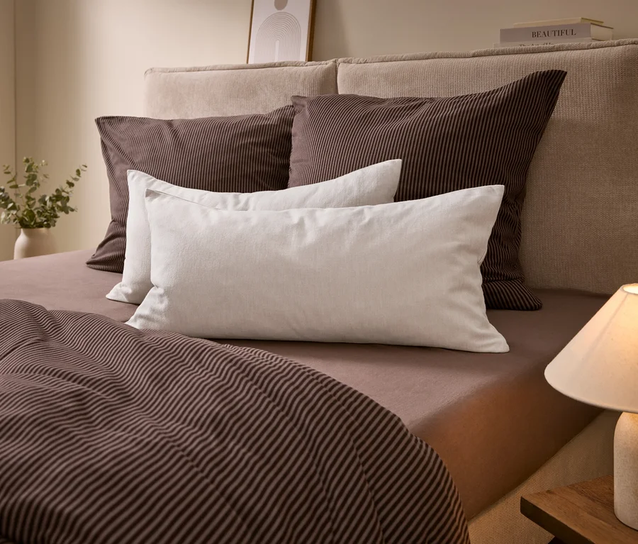 Ein Bett mit beigem Kopfteil, braun gestreifter Bettwäsche und weißen Kissen. Eine Lampe steht auf einem Nachttisch, eine Vase mit Grünpflanzen links.
