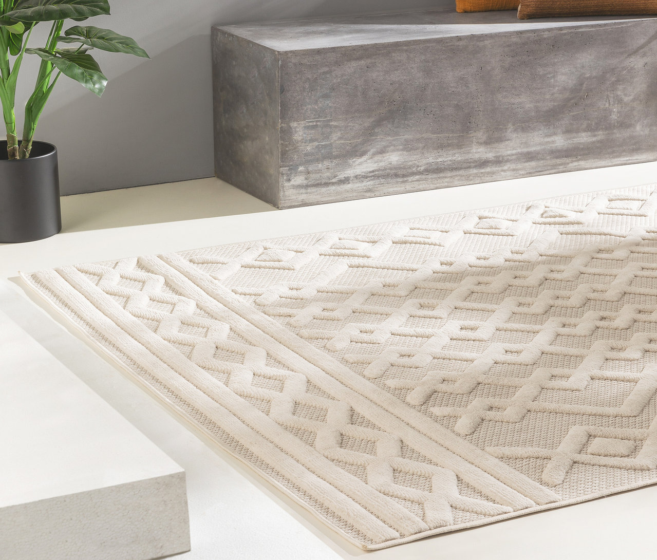 Ein beigefarbener benuta In-und-Outdoor-Teppich »Bonte« mit erhabenem geometrischem Muster liegt auf hellem Boden vor einer grauen Betonbank und einer Topfpflanze.
