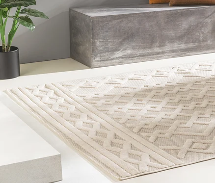 Ein beigefarbener benuta In-und-Outdoor-Teppich »Bonte« mit erhabenem geometrischem Muster liegt auf hellem Boden vor einer grauen Betonbank und einer Topfpflanze.
