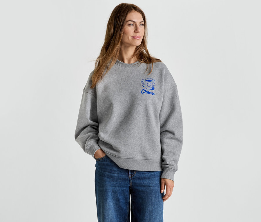 Frau mit grauem Pullover mit blauem 'Cheers'-Aufdruck und Jeans.