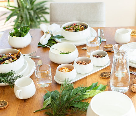 Ein Holztisch ist mit weißen Geschirrteilen des Mäser Teller-Sets „Taya“ gedeckt, darunter Schüsseln mit Salat, Nüssen und Saucen. Gläser, eine Karaffe, Tannenzweige und getrocknete Orangenscheiben ergänzen die Dekoration.